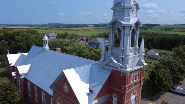 Cinematic drone moving toward the steeple of the historic red-brick church and adjacent cemetery in Saint-Anaclet-de-Lessard village. Cultural heritage of Quebec, Canada.
