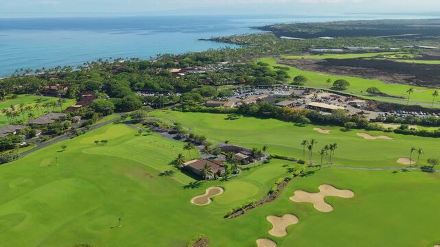 Big Island Hawaii golf resort seen from a drone with green fairways sand bunkers palm trees and a clubhouse beside turquoise ocean coastline lava fields and homes under bright tropical sun.
