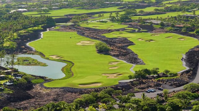 Aerial drone view of luxury golf course on Big Island Hawaii with emerald fairways sand bunkers shimmering pond black lava fields palm trees and resort villas under bright tropical sunlight.