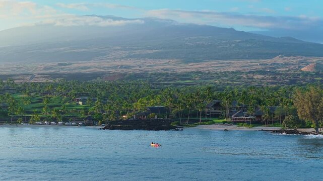 Drone shot of Big Island Hawaii coastline at sunrise with two kayakers paddling calm blue ocean water past palm trees luxury resort villas golden lava fields and volcano slopes beyond.