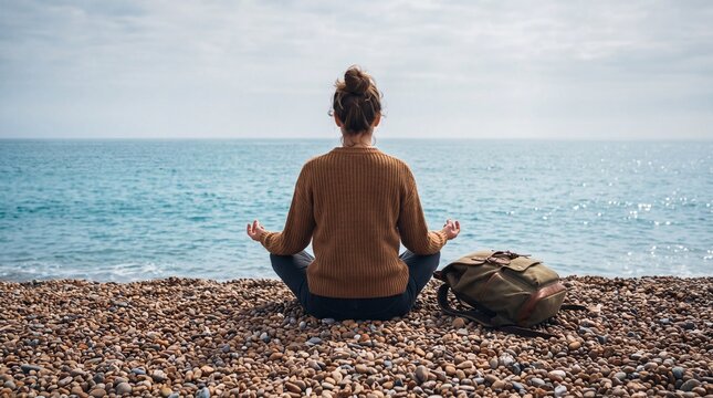 Rear view of a young woman sitting in meditation pose on a pebble beach, enjoying the peaceful ocean view during a travel trip with her backpack.