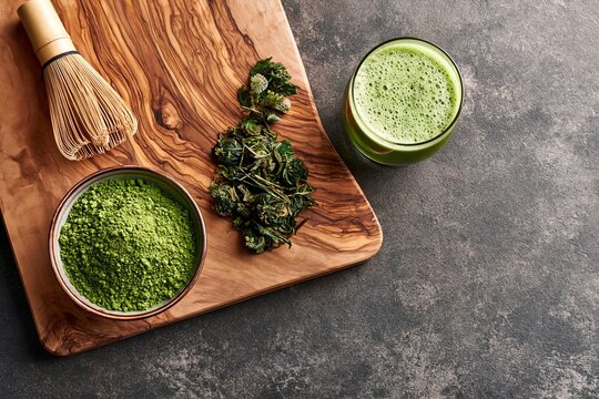 Matcha green tea powder in bowl with bamboo whisk and drink in glass on board