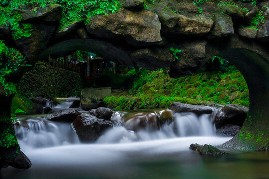 Water flows gently under mossy stone archway. Rocks gleam as water cascades over them softly. Greenery thrives where damp shadows linger. The stream whispers through nature's quiet embrace