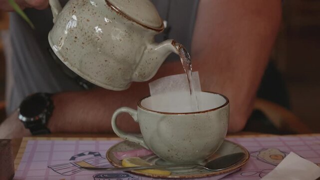 Individual pours hot water from a ceramic teapot into a white cup containing a teabag, placed on a colorful table with a patterned placemat