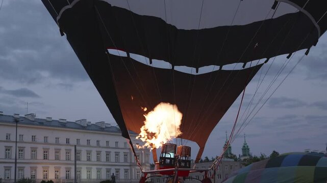 Hot air balloon fills with air as flames burst from the burner, highlighting the colorful fabric and urban architecture during dusk