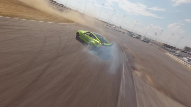 High-speed FPV drone following a green drift car around a track with dust in the air and smoky tires