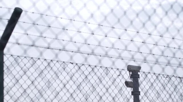 Low angle tilt up video showing modern surveillance cameras mounted behind a chain link fence topped with razor barbed wire. High security, protection, and privacy concept.