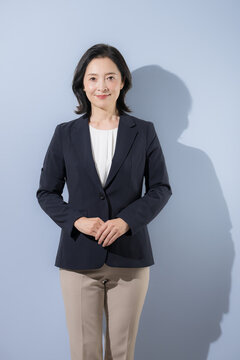 [水色背景]スーツ姿で正面を向いて両手を重ねる立つ日本人ミドル女性、信頼感のある50代の女性ビジネスパーソンのポートレートカメラ目線笑顔