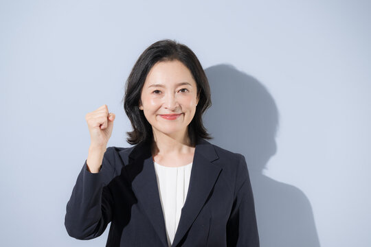 [水色背景]笑顔の日本人ミドル女性、スーツ姿でガッツポーズで元気な明るく前向きな50代の女性ビジネスパーソンのクローズアップ