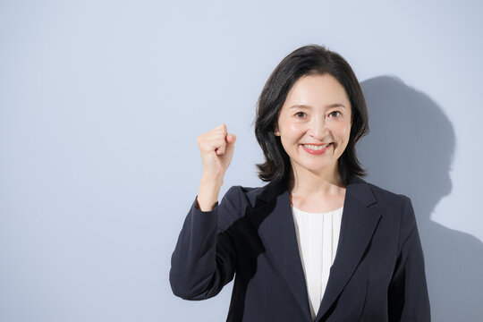 [水色背景]笑顔の日本人ミドル女性、スーツ姿でガッツポーズで元気な明るく前向きな50代の女性ビジネスパーソンのクローズアップ