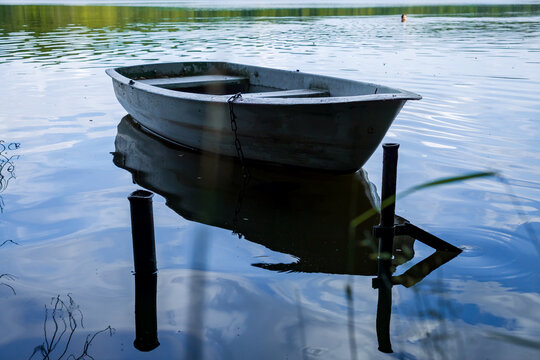 An empty metal rowboat moored on a calm forest lake, with reflections on the water and a distant swimmer blurred in the background. Peaceful summer scene representing solitude and relaxation.