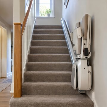 Home stairlift seat folded against the wall on a carpeted staircase with soft daylight from the stairwell window