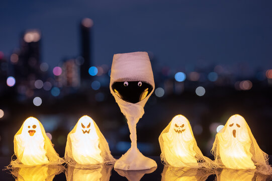 Halloween mummy red wine glass with glowing ghost lights against a city skyline at night.