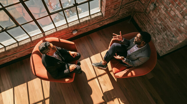 Business Dialogue: Two professionals engaged in a deep conversation, seated in modern chairs by a large window, bathed in natural light.