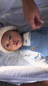 Vertical Screen: African American newborn baby in a knitted bonnet and blue knit romper lies awake in a soft bassinet while a parent hand gently touches the tiny fingers indoors.