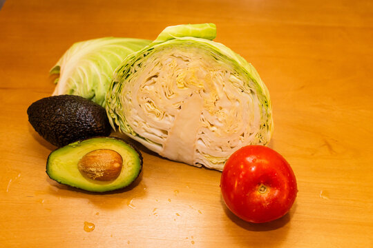Vista superior de ingredientes frescos para ensalada: repollo verde cortado, un tomate h&uacute;medo y aguacate con semilla sobre una tabla de madera.