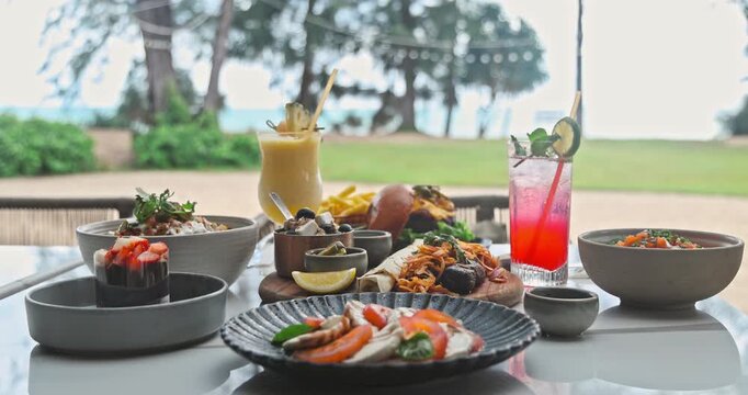 Gourmet food and colorful cocktails on a dining table at a tropical beach resort. Fresh caprese salad, burger, and refreshing drinks served outdoors with a scenic ocean view. Summer vacation lifestyle