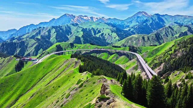 Aerial view of a curved highway viaduct winding through a lush green mountain valley in Xinjiang, China
