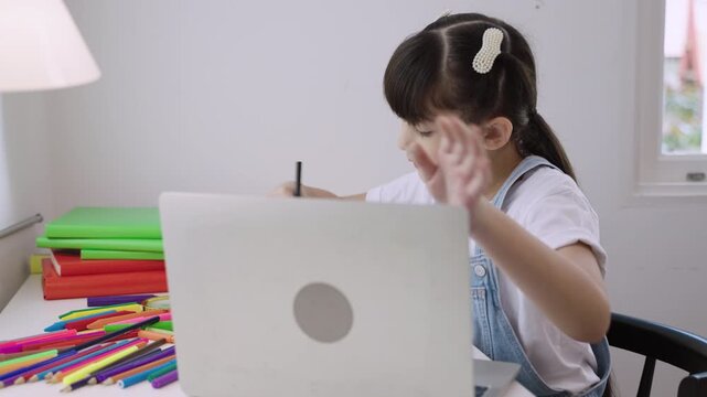 Cute Asian girl student sitting at desk using a laptop computer for studying online learning via video calling with remote teacher in a home school. Homeschooling education and new normal concept. 