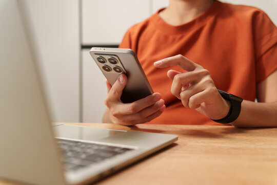 Asian Woman using smartphone beside a laptop at a desk, representing mobile communication, online browsing, multitasking, digital technology, remote work and connected modern lifestyle.