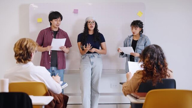 Three university students deliver a team presentation using notes and a tablet before classmates in a modern Latino college classroom