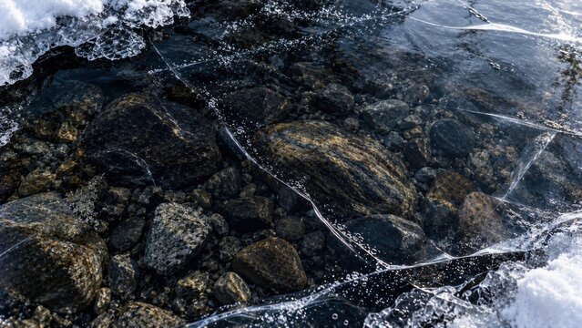 Lake ice fracture above submerged stones, radiating crack lines, transparent black ice and trapped white bubbles. Winter texture background for design. For climate media and seasonal publishing.