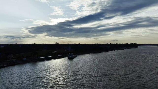 Aerial view of a dark tree-lined shoreline and calm rippling water beneath dramatic clouds at sunset, with small docks and homes along the coast