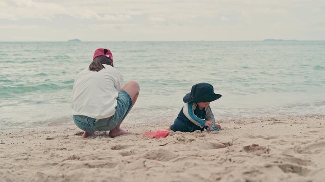 Happy mother and adorable baby boy sitting on sandy beach together during summer vacation, slow motion