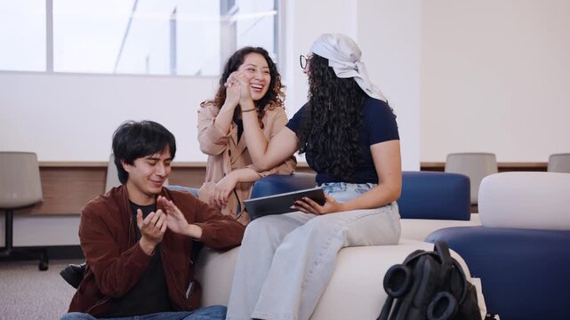 Two Latina university friends exchange a playful high five while a male classmate claps and laughs during a campus study break