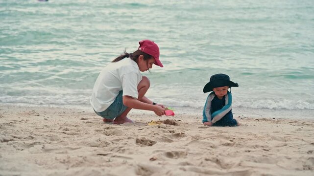Happy mother and adorable baby boy sitting on sandy beach together during summer vacation, slow motion