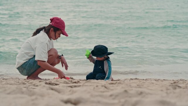 Happy mother and adorable baby boy sitting on sandy beach together during summer vacation, slow motion