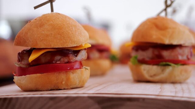 Close-up of freshly prepared mini cheeseburgers with meat patties, cheese, tomato and greens served on a wooden board. Appetizer and party food concept.
