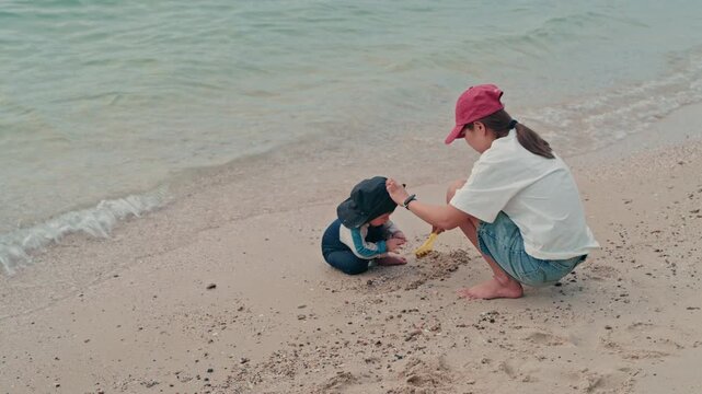 Happy mother and adorable baby boy sitting on sandy beach together during summer vacation, slow motion