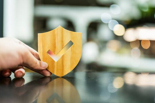 Verified protection and security concept, hand holding wooden shield with checkmark cutout on reflective table against modern bokeh background, symbolizing safety, trust, and quality guarantee.