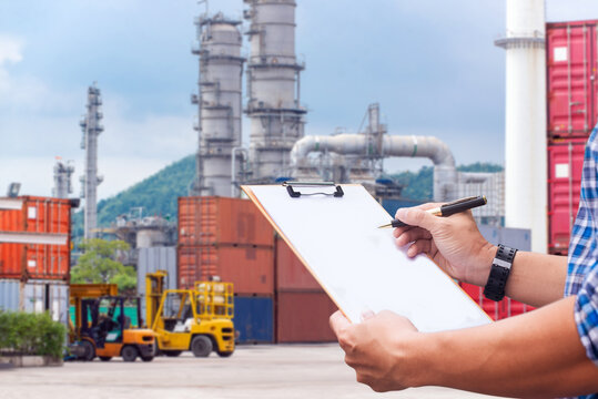 Logistics inspector writing on clipboard at cargo container terminal with oil refinery background. Human-created, not AI.