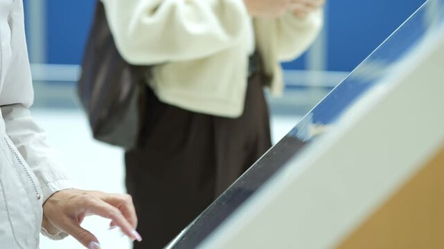 Person points finger at touch screen kiosk. Second person stands nearby holding towel. Background shows indoor public space with blue walls. Focused shot on hand interaction with modern technology
