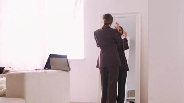 Business dressing. Workday preparing. Stylish female in modern brown trouser suit using mirror checking outfit before leaving home.
