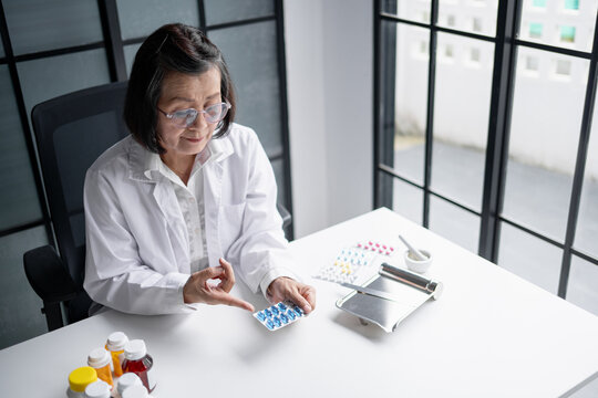 Asian senior female pharmacist explaining prescription medication while holding blister pack at pharmacy counter.