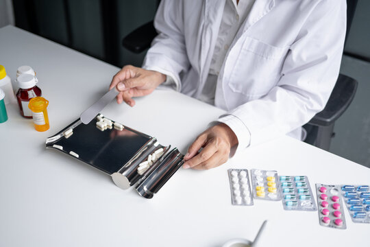 Asian senior female pharmacist counting and arranging prescription pills on stainless steel counting tray with spatula at pharmacy counter.