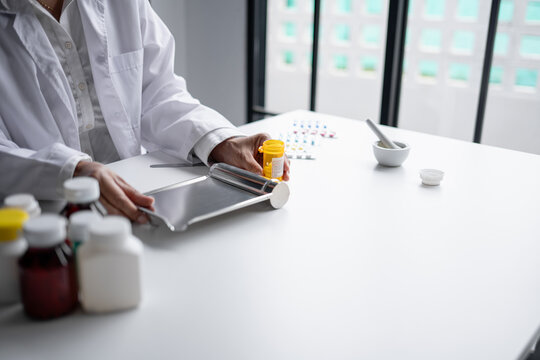 Asian senior female pharmacist counting and arranging prescription pills on stainless steel counting tray with spatula at pharmacy counter.