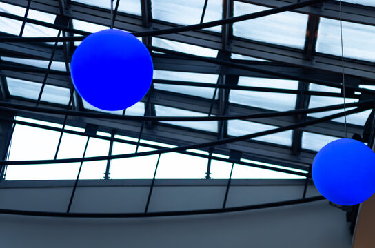 Glowing blue spherical lanterns hanging from the ceiling structure inside the Batory Shopping Center in Gdynia, Poland. Modern interior design featuring a glass roof with dark steel structural beams.