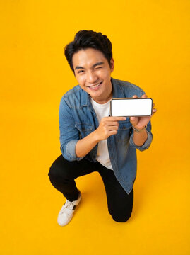 Young man holding blank mobile phone screen while winking at camera wearing casual denim shirt against bright vibrant yellow background for digital marketing advertising campaign