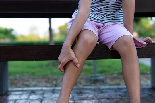 Close-up of young Asian girl sitting on park bench scratching itchy calf and insect bites on leg