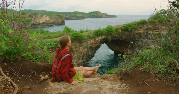 Woman sits enjoying peaceful natural view of Broken Beach rock arch cliffside landscape Nusa Penida Bali