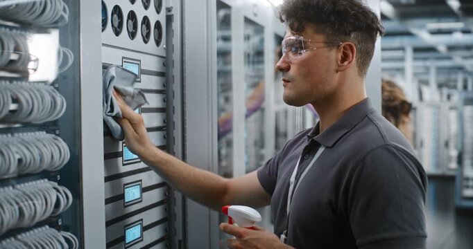 Network Engineer Wearing Protective Glasses Holds Detergent Spray Bottle while Wiping Down Metallic Server Rack Modules. Concept of Hardware Maintenance, Anti Static Cleaning, Data Center Cleaning.