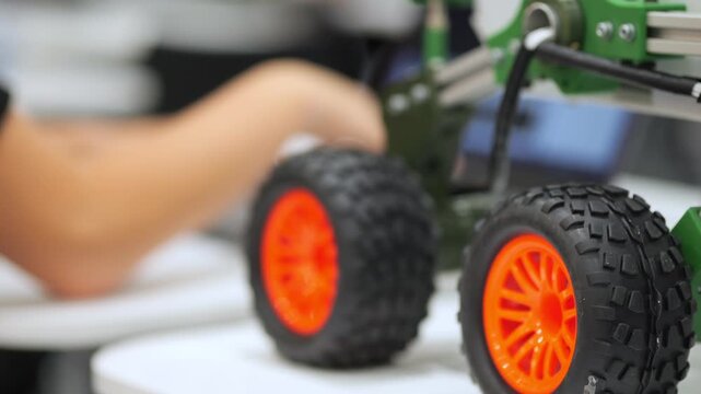 Close-up of orange wheel toy car. Blurred hands work nearby. Green robotic vehicle detail. Tech workshop environment shown