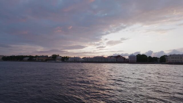 Static wide shot of historic riverside embankment with neoclassical architecture silhouetted against dramatic twilight sky, natural golden hour lighting reflecting on calm water.