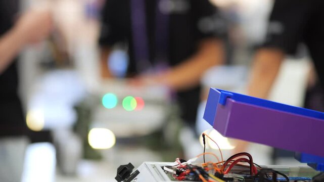 Close-up of electronic circuit board with wires and glowing lights. Background shows blurred people at tech event. Focus on technical components and active devices