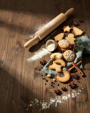 An overhead flat lay of festive holiday cookies arranged on a rustic dark wooden table with a wooden rolling pin, dusted flour, cinnamon sticks, star anise, and evergreen sprigs for a cozy vibe