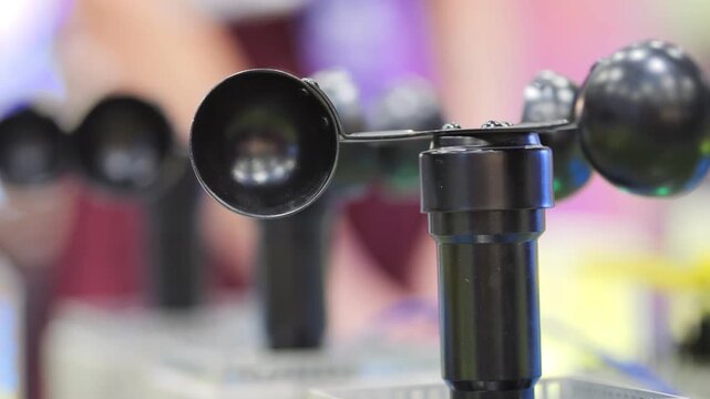Close-up of black anemometer with rotating cups. Multiple similar instruments visible in background. Blurred indoor setting suggests science fair or exhibition